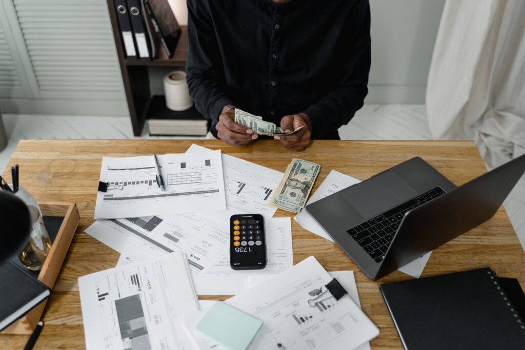 Person counting cash at paper covered desk with phone open to calculator