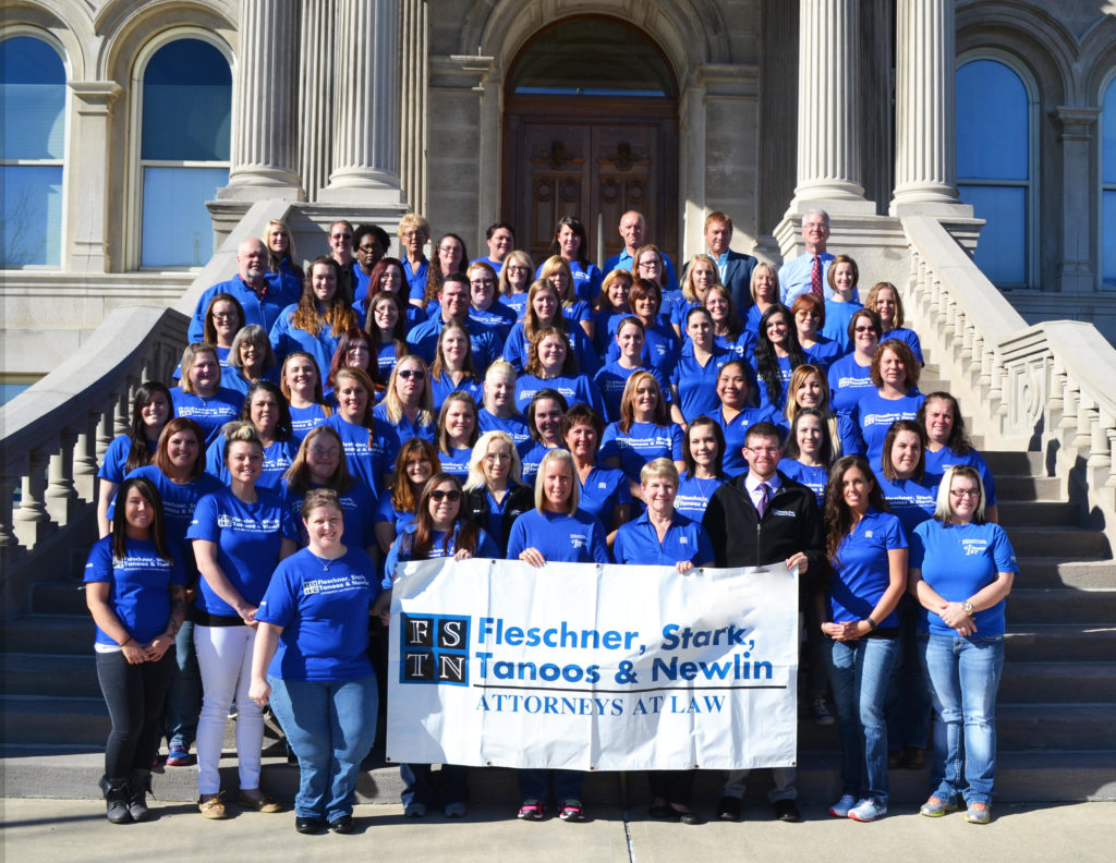large group of people wearing blue t-shirt posing for photo while standing on stairs and holding a sign that reads: "Flescher, Stark, tanoos & Newlin"