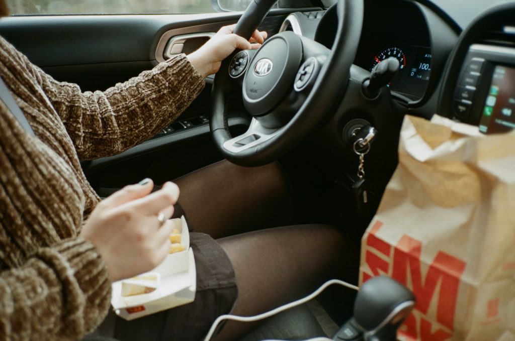 A young woman has fast food in her lap while she driver har car.